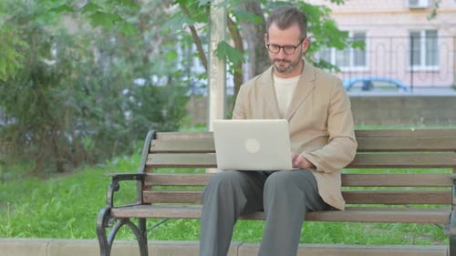 Man Works on Laptop on Bench in Park