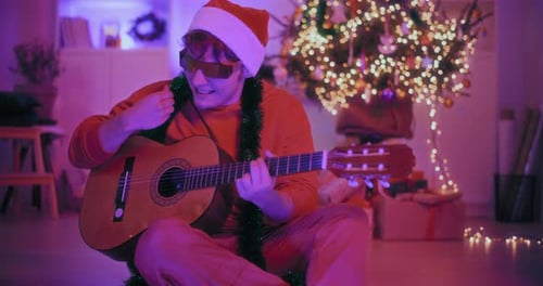 Excited Man Playing Guitar While Sitting on Floor at Illuminated Home During Christmas