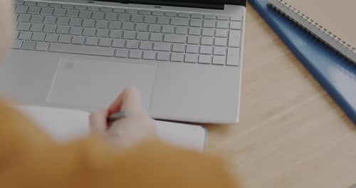 Nahaufnahme einer weiblichen Hand mit Laptop, die Informationen in ein Notizbuch am Schreibtisch tippt und schreibt