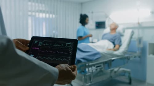 In the Hospital Doctor Holds Tablet Computer Showing Vital Signs of a Senior Patient Lying in Bed.