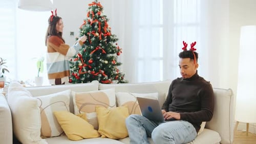 Multiracial Couple Celebrates Christmas As Woman Decorates a Festive Tree While Man Works on Laptop