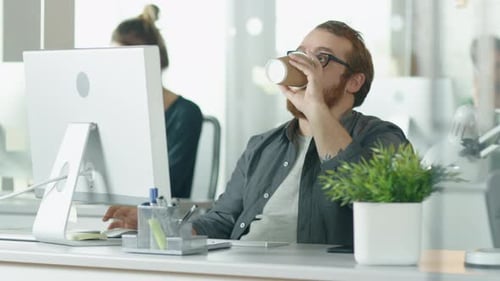 Bärtiger junger Mann arbeitet an seinem Desktop-Computer und trinkt Kaffee. Im Hintergrund arbeitet das Mädchen an ihrem Schreibtisch.