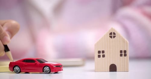Close-up of a hand holding a pencil and writing something on a yellow paper, behind a red toy car th