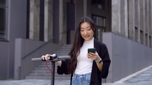 Asian woman using her smartphone and walking with personal e-scooter near modern building