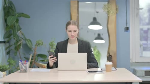 Woman Uses Smartphone at Desk with Laptop