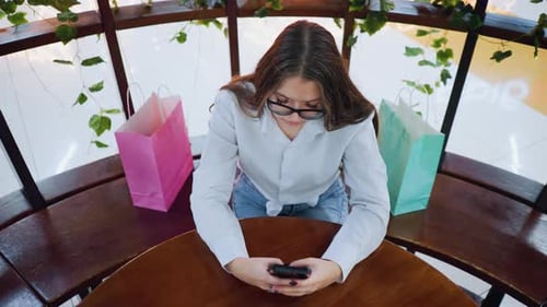 Overhead View of Woman Chatting with Friend Over Phone with Shopping Bags at Round Table