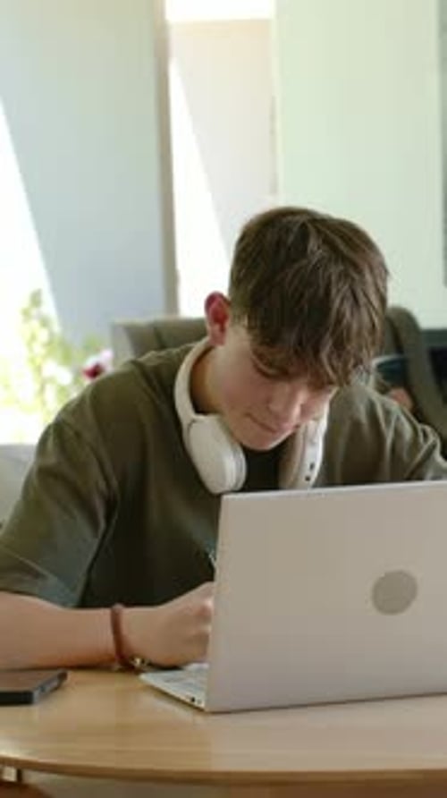Vertical video: Glancing at laptop, student writing notes for homework at home table with notebook