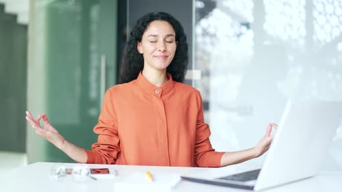 Young female employee is meditating with her eyes closed sitting at a workplace in office.
