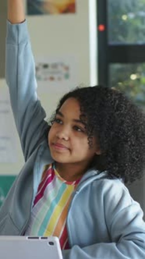 Vertical video: Raising student in blue hoodie lifting right arm answering teacher at desk, tablet