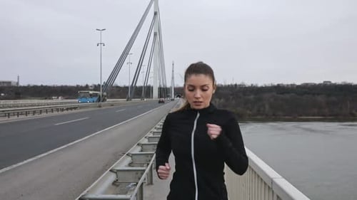 Young woman in black sports outfit running on the bridge in the city during day.