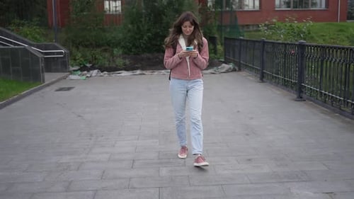 Young Woman is Walking Down a Paved Path While Looking at Her Smartphone She is Smiling and Seems to