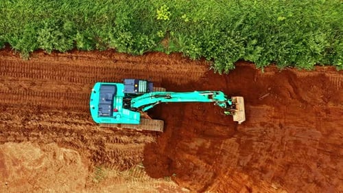 Aerial view of bright blue excavator leveling red soil near green overgrowth