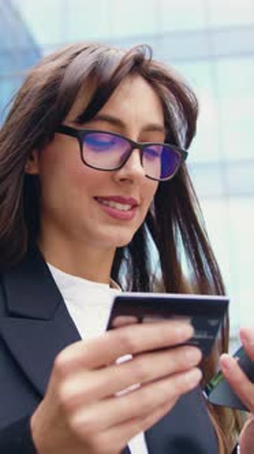 Attractive Caucasian Woman Standing Outdoors in a Modern City Using a Smartphone and Credit Card to