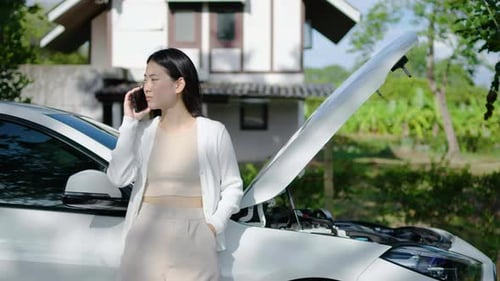 Anxious young asian woman standing near a broken car with popped up hood calling talking on phone.