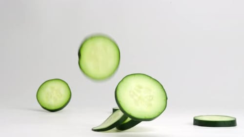 Fresh green cucumber coins falling onto table, bouncing, flipping and settling into a pile in slow m