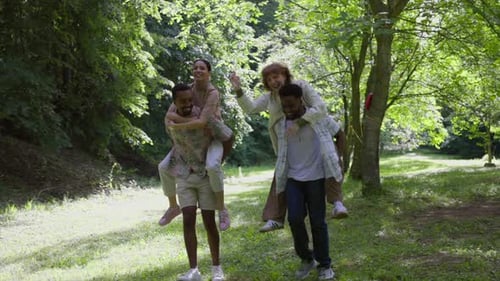 Two men giving piggyback rides to their girlfriends in a sunny park, surrounded by nature