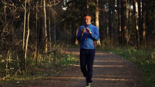 Young Man in Sportswear Walking Along a Forest Path Looking at His Smartphone to Check His Training