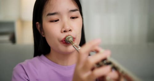 A young woman practices playing the trumpet while sitting on a sofa in a cozy living room.