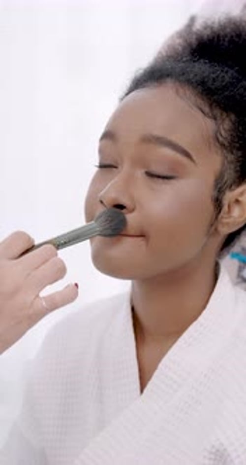 Jeune adolescente afro-américaine assise dans un salon de beauté souriant brillamment pendant qu'elle est maquilleuse...