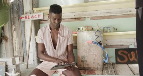 Happy african american man using tablet and smiling at surfboard rental beach shack, slow motion