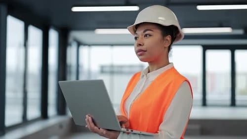 Professional Female Architect in Hard Hat Reviews Plans on Laptop in Office Space