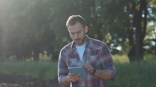 Ernsthafter konzentrierter junger Mann benutzt und tippt auf Tablet im Freien Landwirt überwacht Ernte in der