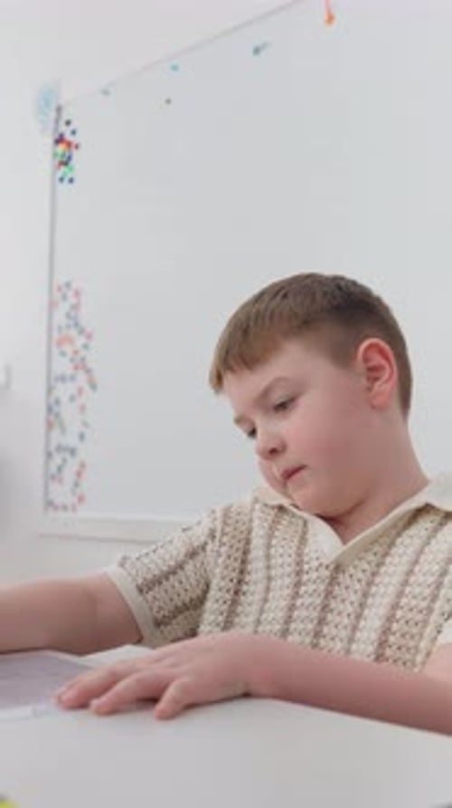 Young Boy Seated at a White Table in a Classroom Looking Down