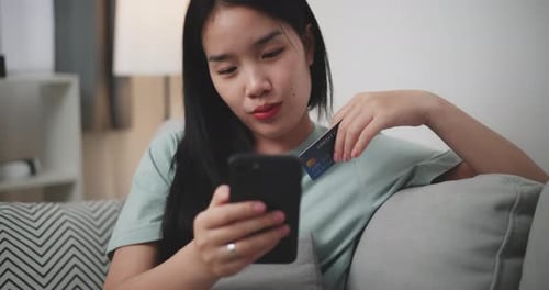 Young woman uses phone and credit card to shop online on sofa at home.