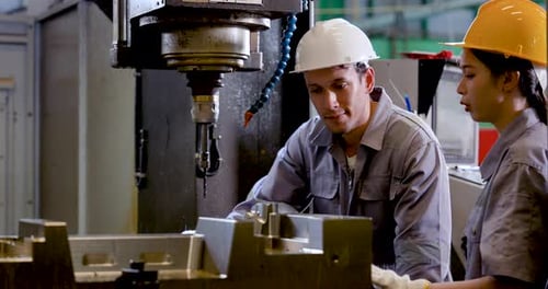 Asian man engineer leans toward CNC fixture while Asian woman technician monitors area, discussing