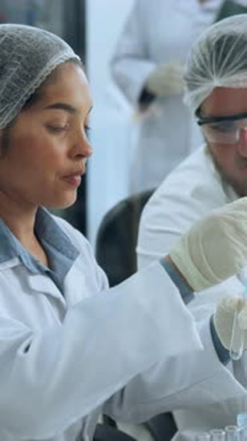 Woman Scientist Pipetting Liquid in Lab, Man in Background