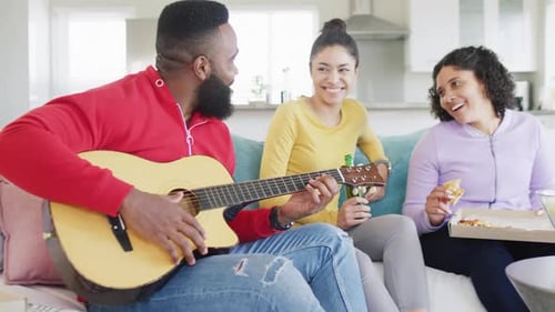 Happy, diverse female and male friends playing guitar and listening at home in slow motion