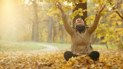 A Girl Sitting on the Ground Scatters Fallen Yellow Leaves and Enjoys the Park