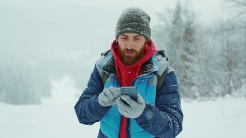 Portrait Young Man Outdoor in Winter Snowy Forest Using Smartphone for Message Online Christmas