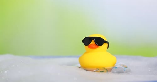Cute rubber duck floating in water with bubbles against blurred green background, closeup