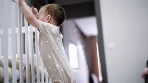 Small kid shakes a crib excitedly. Baby boy plays in his room. Low angle view.