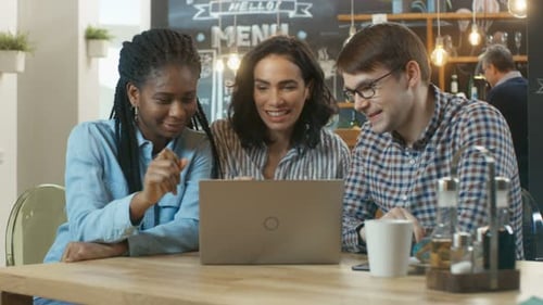 Three Young People Have Fun in the Coffee Shop, They Watch Something on a Laptop and Talk. In the B