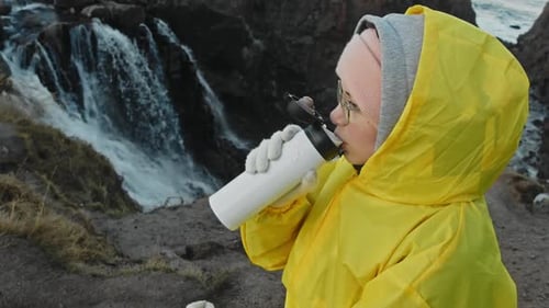 Woman Drinks Water by a Waterfall in Yellow Raincoat