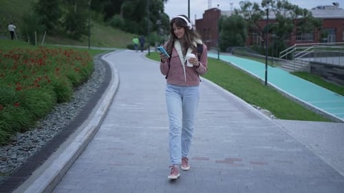 Young Woman Walks in the Park While Listening to Music on Her Headphones and Drinking a Coffee She
