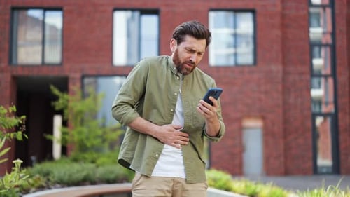 Upset Man Feeling Stomach Pain While Using Phone Near Apartment Building