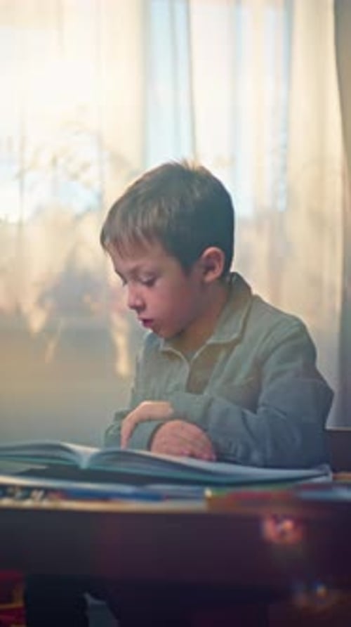 Boy Reads Book at Table by Window
