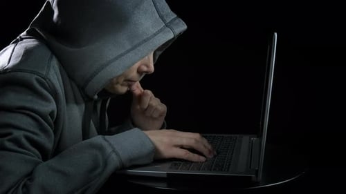 Hooded Figure Typing on Laptop in Dark Room