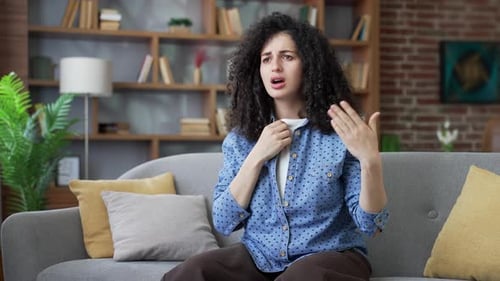 Dissatisfied young beautiful female is hot while sitting on sofa in living room at home.