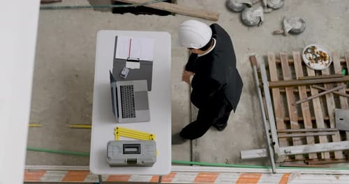Asian construction inspector crouches over floor plan near building material, checking survey
