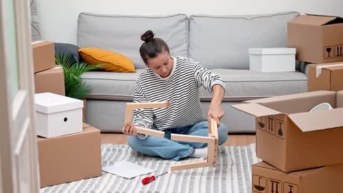 Woman Assembling Shelf Amidst Moving Carton Boxes and Furniture Making Her New Place Feel Like Home