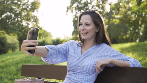 A Young Caucasian Woman Talks to a Smartphone with a Smile As She Sits on a Bench in a Park
