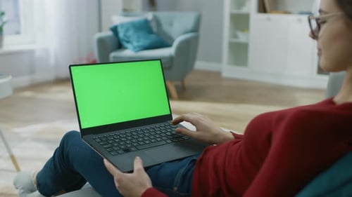 Young Woman at Home Works on a Laptop Computer with Green Mock-up Screen. She's Sitting On a Couch
