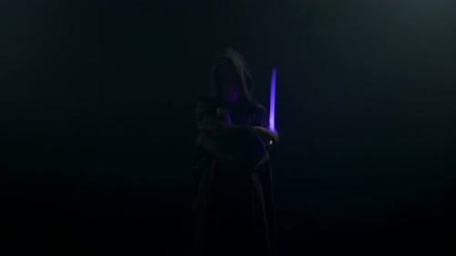 Young Man in Jedi Cosplay Costume with Lightsaber Battle on Black Background in Smoke and Rain Slow