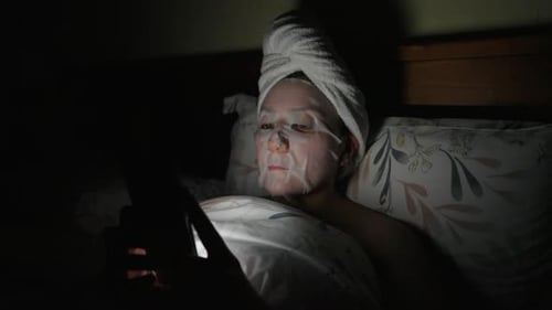 Woman in Bed with Face Mask on Phone