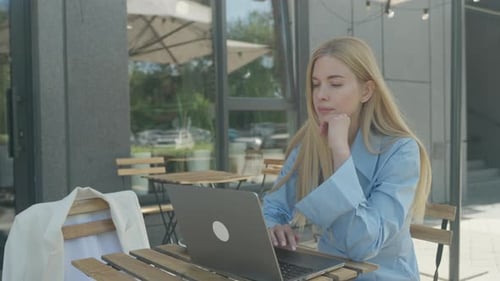 Smiling Blonde Woman Working On Laptop And Fixing Hair At Outdoor Cafe