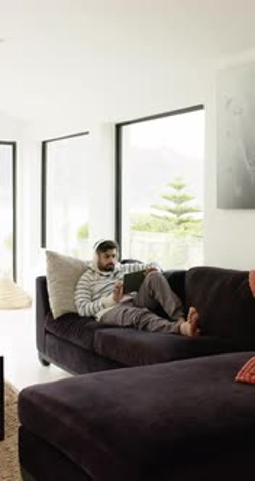Vidéo verticale : homme indien allongé sur un canapé, tapotant une tablette avec un casque dans un salon lumineux